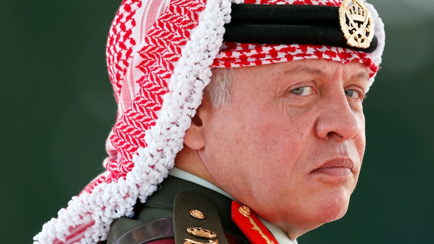 King Abdullah II looks over his shoulder at the camera in a close-up shot. His appearance is stern he wears military attire.