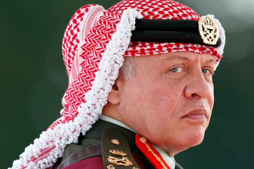 King Abdullah II looks over his shoulder at the camera in a close-up.  His appearance is severe, he wears military attire.