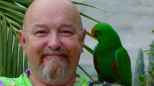 A smiling bald man with a goatee wears a bright shirt and a parrot on his shoulder.