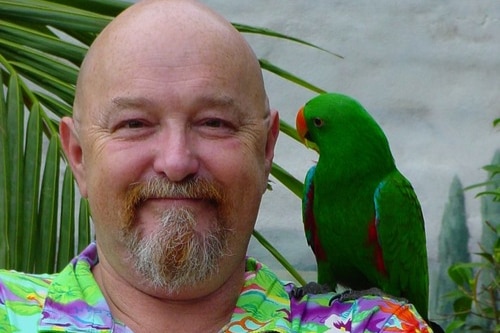 A bald man with a parrot on his shoulder smiles at the camera. 
