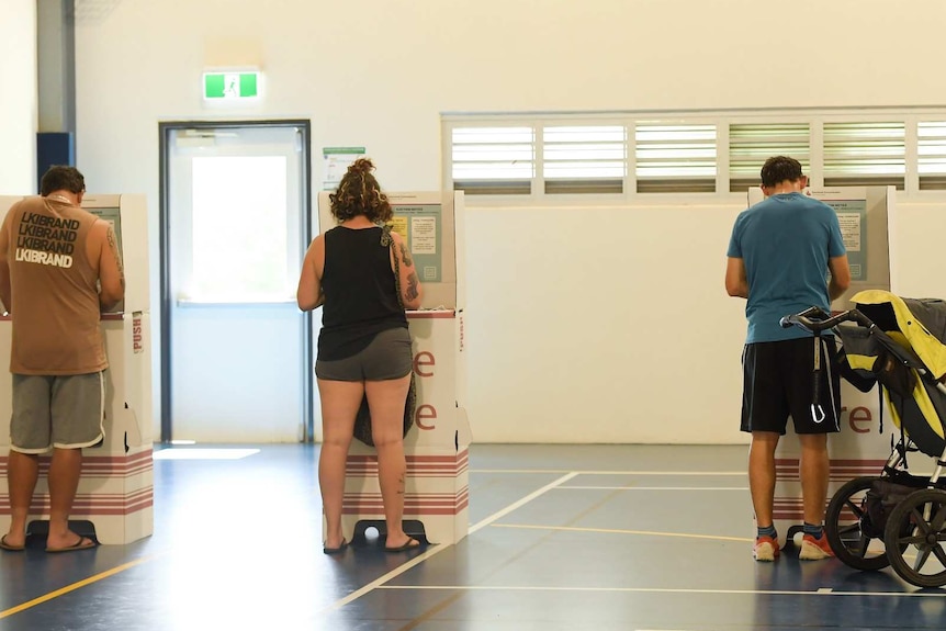 Three people vote at ballot boxes on a basketball court.