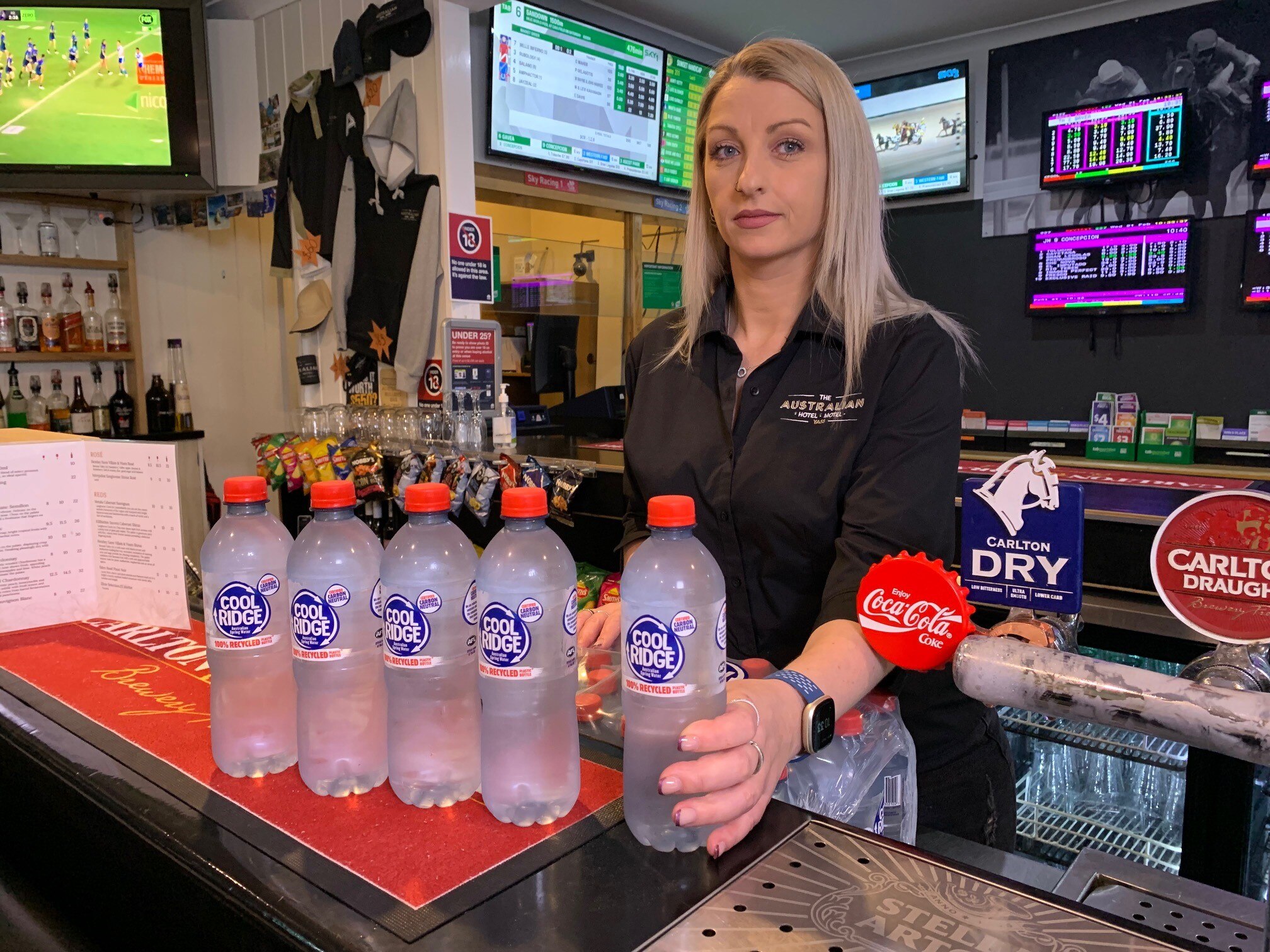 A woman stands behind a pub bar holding five bottles of water.