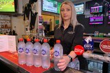 A woman stands behind a pub bar holding five bottles of water.