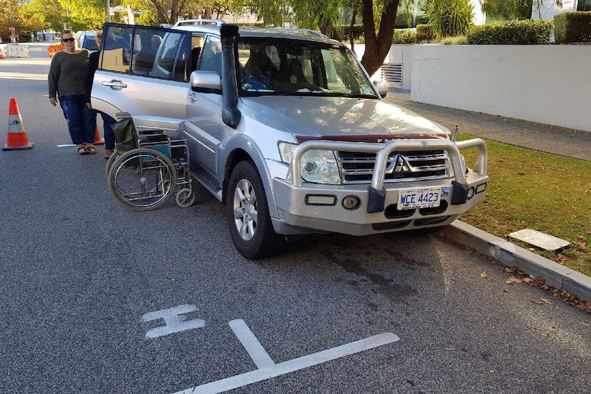 A silver four wheel drive car with a wheelchair next to it in a parking spot on the roadside.