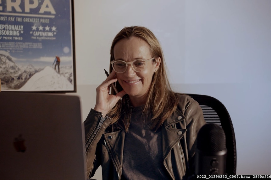 A white woman with long honey blonde hair wears a black leather jacket and pale glasses and sits at a screen smiling.