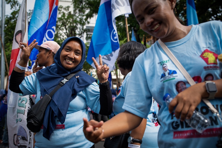 Women in blue shirts with flags behind smile and offer the peace sign to the camera
