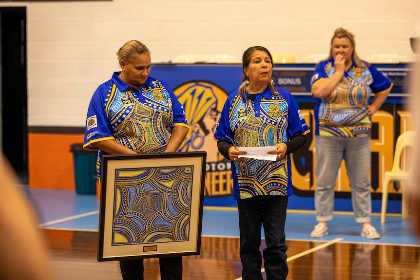 Three ladies standing in matching shirts, one holding a painting and another talking while holding a piece of paper.   