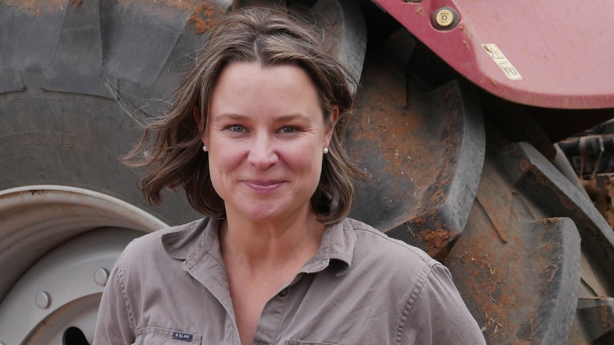a woman with short borwn hair and a brown rb sellars shirt stares into the camera, a tractor wheel is behind her
