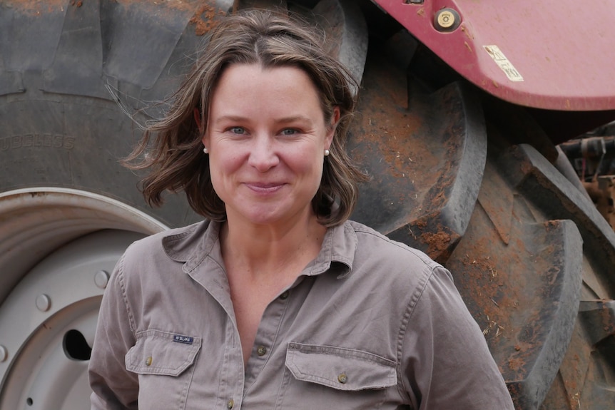 a woman with short borwn hair and a brown rb sellars shirt stares into the camera, a tractor wheel is behind her