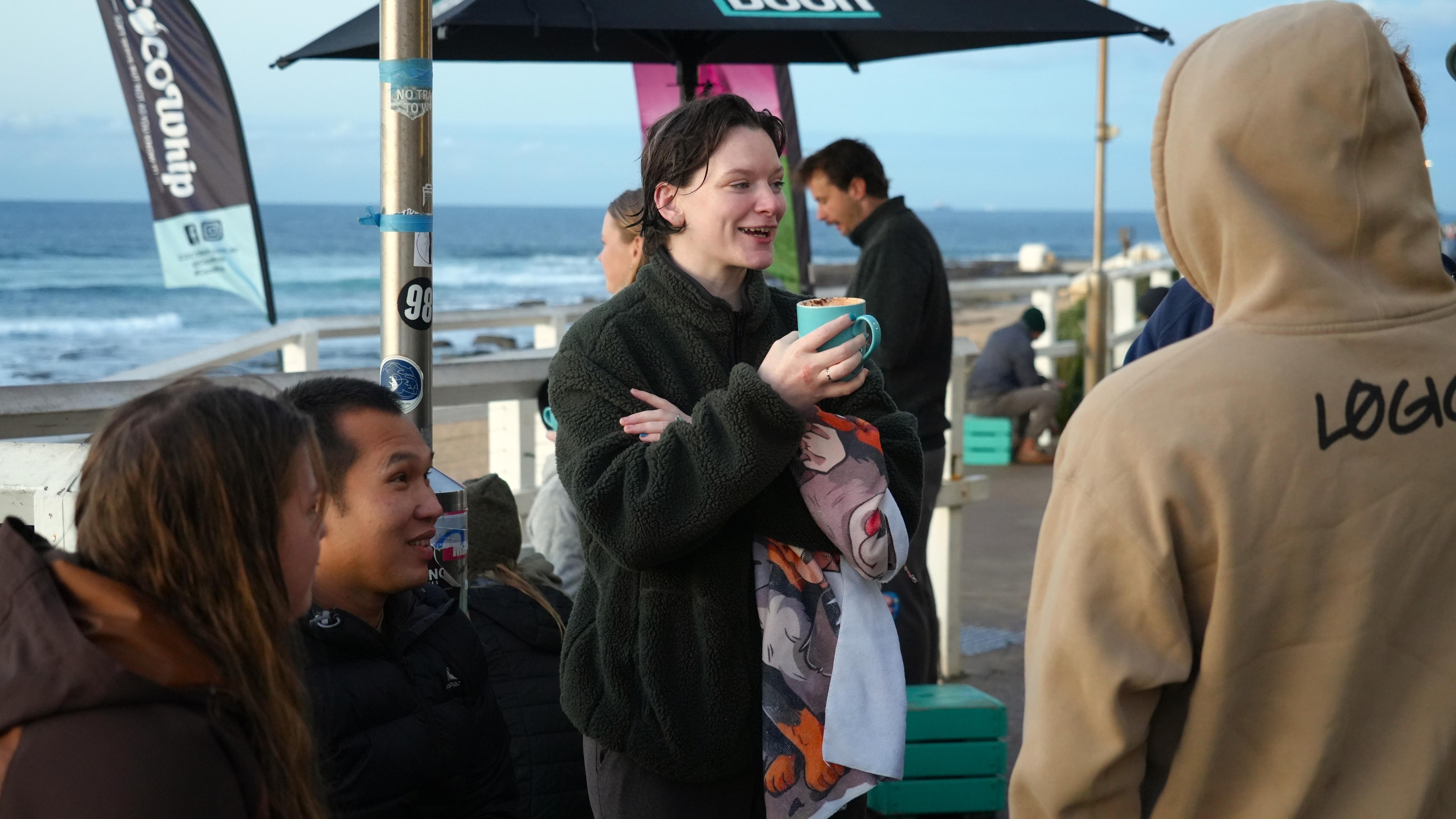 A woman stands with a coffee cup in front of the beach.