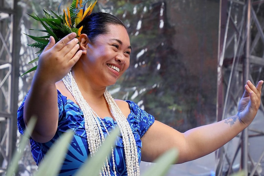 Green leaves in the foreground with a woman in a blue dress and white necklace dancing and smiling