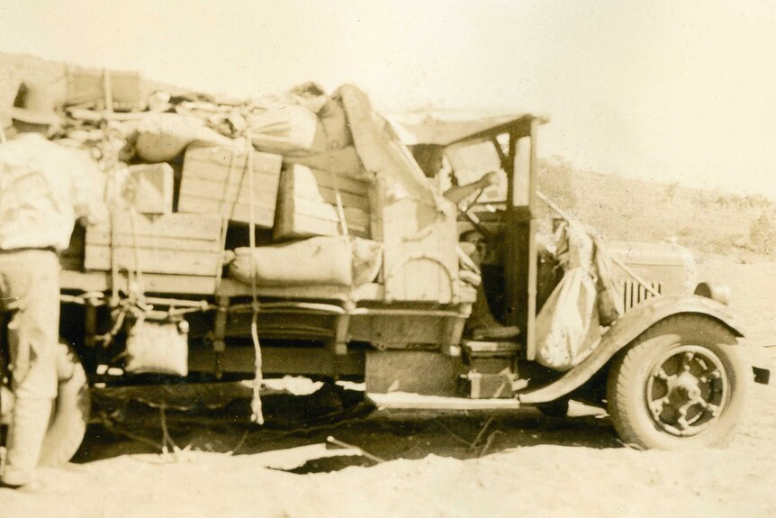 An old 1920s truck laden with crates is stuck in sand in the desert in a sepia-toned photo.