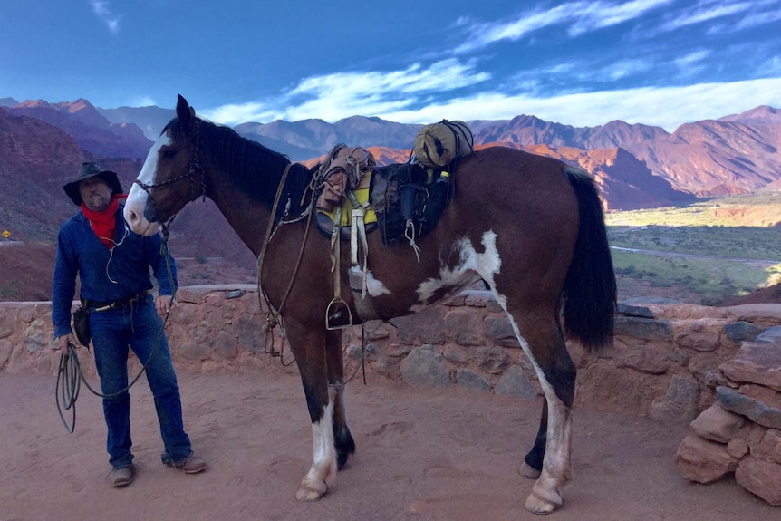 A man in a cowboy hat and riding gear stands by his brown and white horse, in front of a wide valley flanked by mountains.