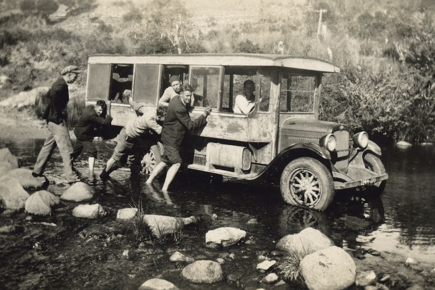 A group of men help push an old bus through a shallow creek.