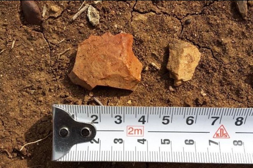 A broken rock and a smaller piece sit on red dirt and are being measured by a tape measure around 4 millimetres