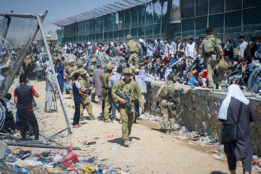 Soldiers in military uniform patrol as swathes of people congregate along a wall