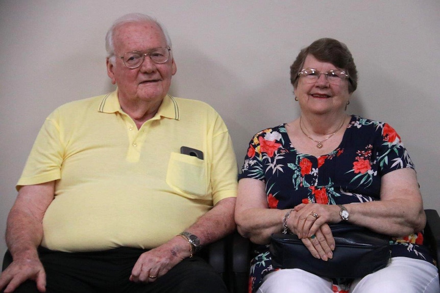 An older couple sit with backs against a wall, the man on the left in a yellow shirt and woman on the right in a black top.