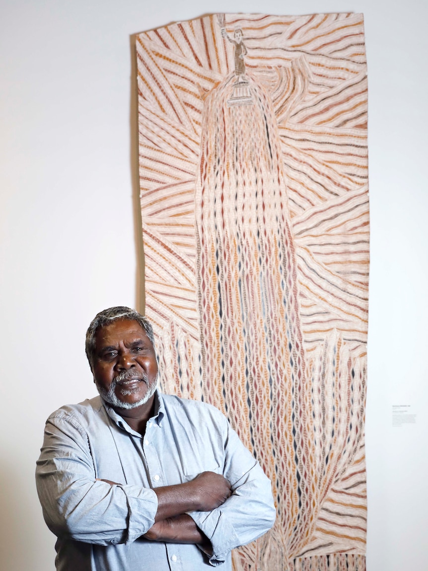 Older man wearing light blue button-down shirt standing in front of white gallery wall hung with long vertical bark painting.