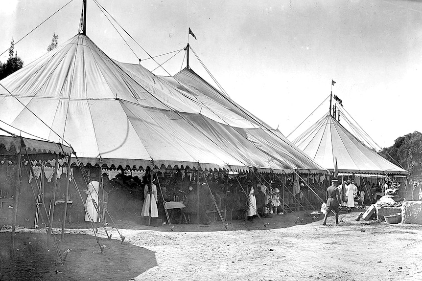 People in early 20th century clothing gather beneath two marquee tents.