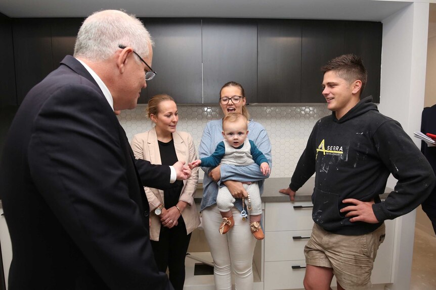 A baby being held by a young woman holds Scott Morrison's finger as they stand in a kitchen