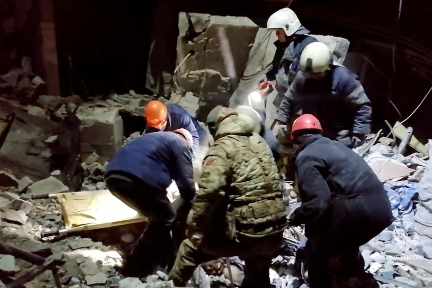A group of men carry a stretcher over a pile of rubble in the dark.