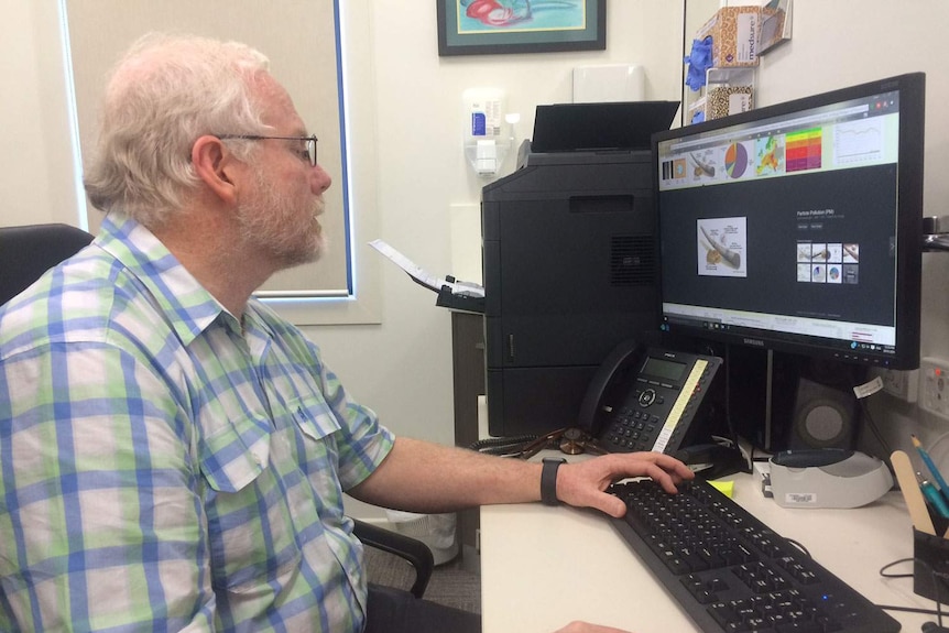 A man in a checked shirt and glasses sits at a computer in his office.