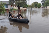 Two children in casual clothes paddle a blue coloured kayak in floodwater in a suburban street.