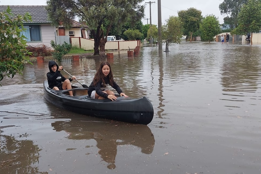 Two children in casual clothes paddle a blue coloured kayak in floodwater in a suburban street.