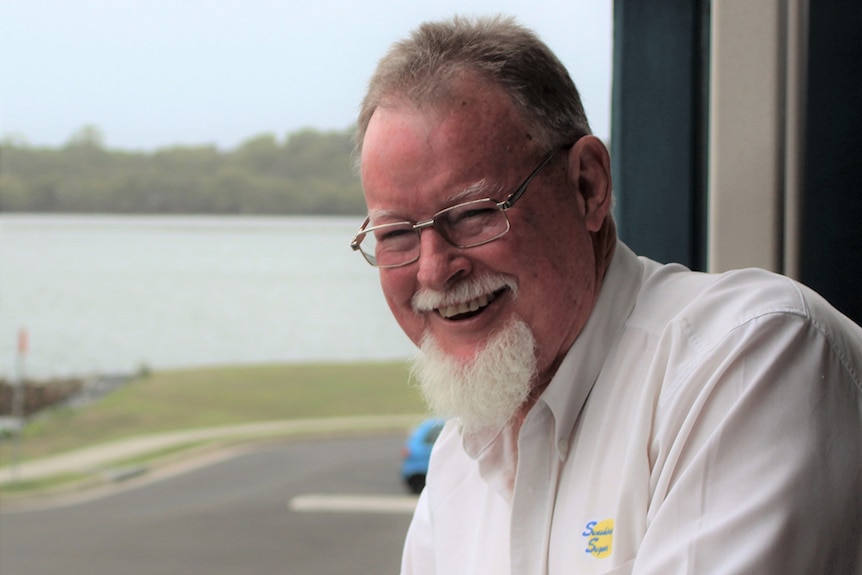 A broadly smiling man with receding hair, a white moustache, goatee wears a white shirt, glasses. River in background.