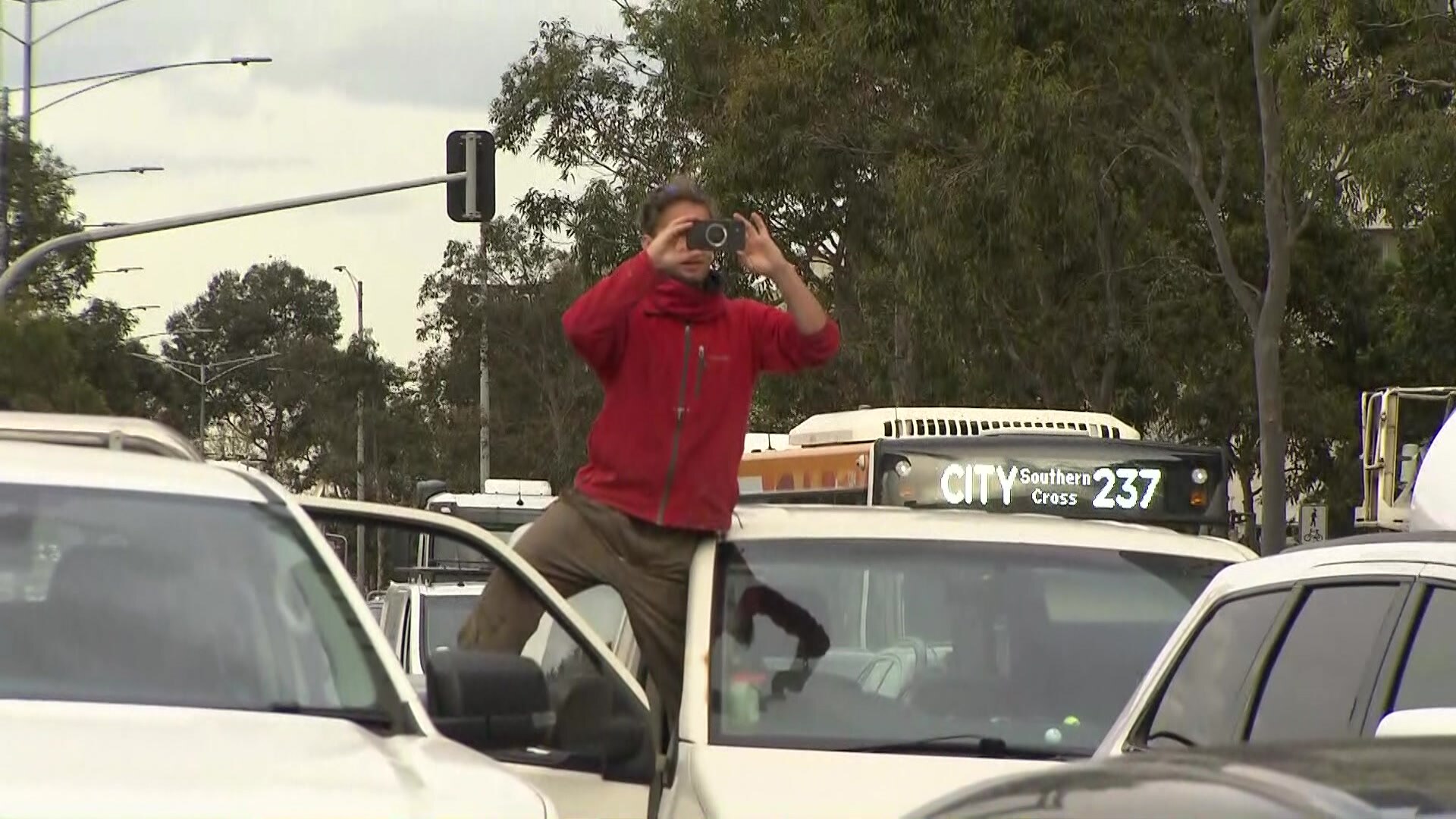 一名男子站在车座上，车门敞开，正在拍照，而他身后则挤满了车辆。