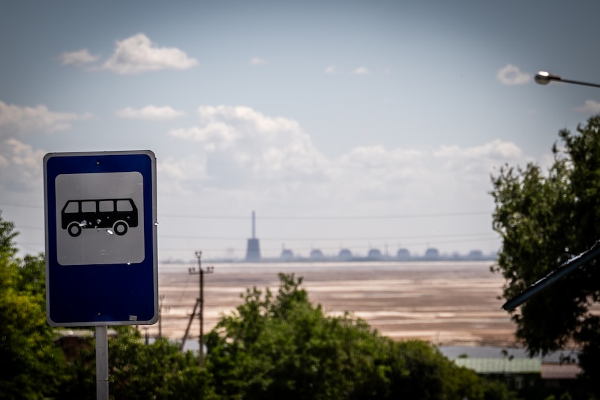 A bus stop sign in the foreground, with a nuclear reactor and buildings visible on the horizon beyond a brown river bed