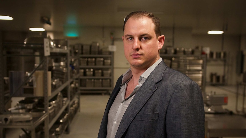 Ben Cook, wearing a suit jacket, looks sternly at the camera in a commercial kitchen.