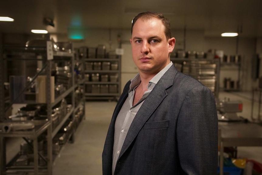 Ben Cook, wearing a suit jacket, looks sternly at the camera in a commercial kitchen.