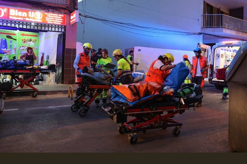 Paramedics push multiple people in stretchers with an ambulance in the background. 