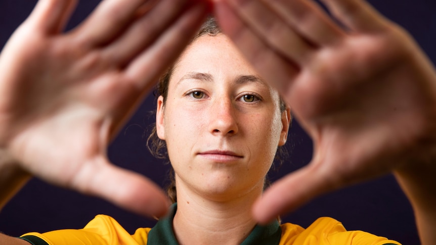 A female soccer player wearing yellow and green holds her hands across a camera lens