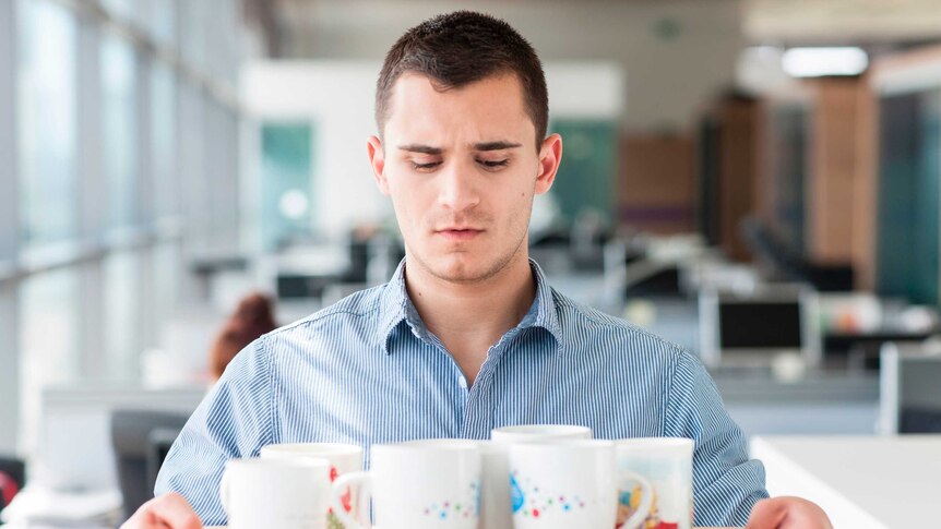 Young man in an office space stares nervously at a full tray of coffee cups he holds in his hands.