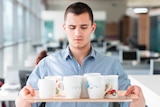Young man in an office space stares nervously at a full tray of coffee cups he holds in his hands.