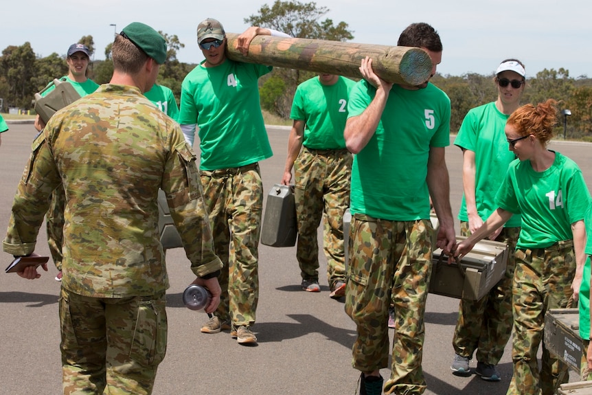 Two male athletes wearing camouflage pants and coloured t-shirts carry a log as a soldier looks on.