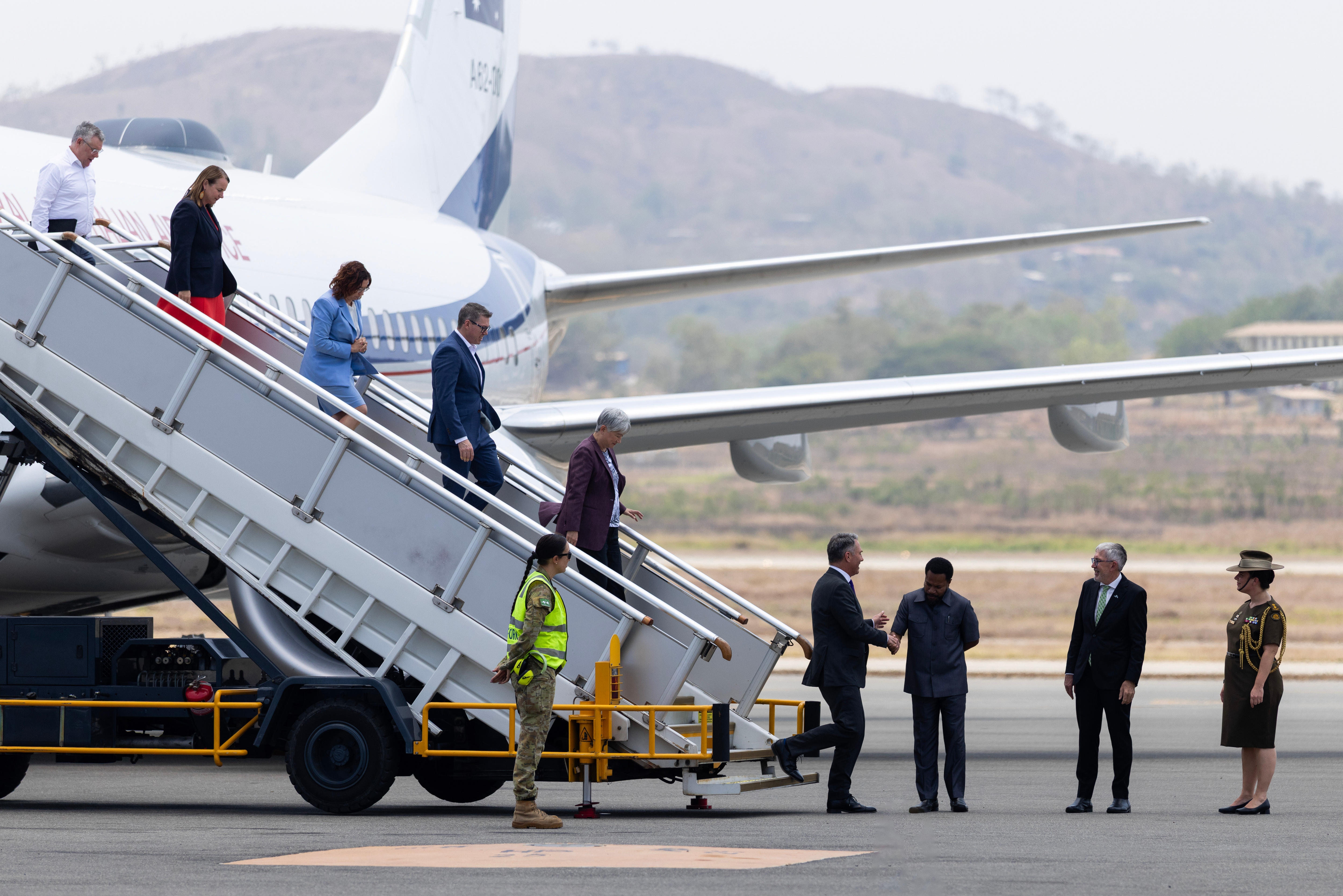 Men and women in suits disembark from a plane and walk down a portable staircase to the airport tarmac. 
