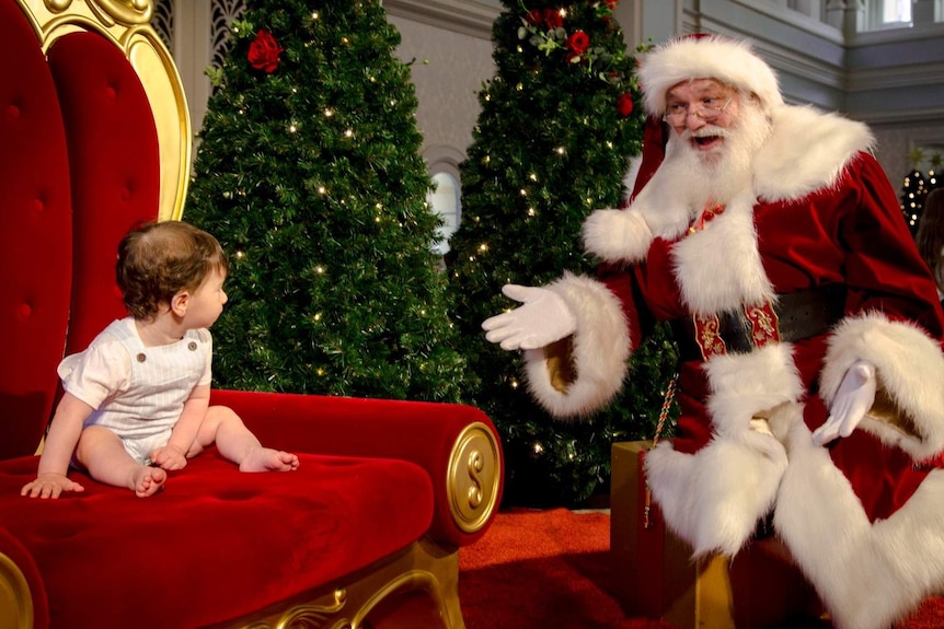 A baby sits on Santa's chair. Santa is to the right of the photo with a surprised look on his face.