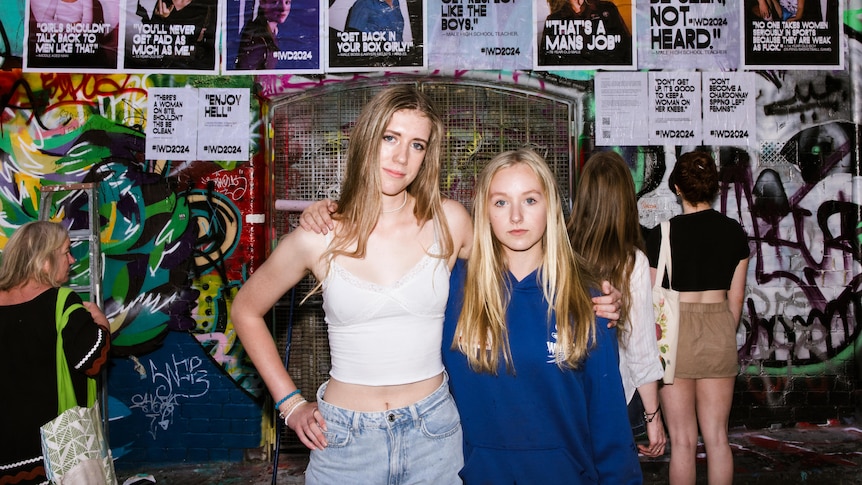 Mia and Eliza are standing with arms around each other, grafitti in background with posters glued to the wall