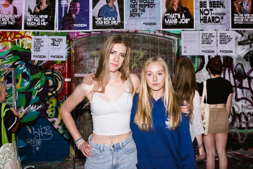 Mia and Eliza are standing with arms around each other, grafitti in background with posters glued to the wall