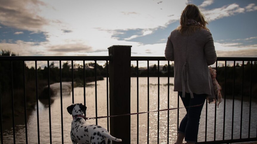 A blonde woman looks out over a river, with her dalmation on a lead beside her.