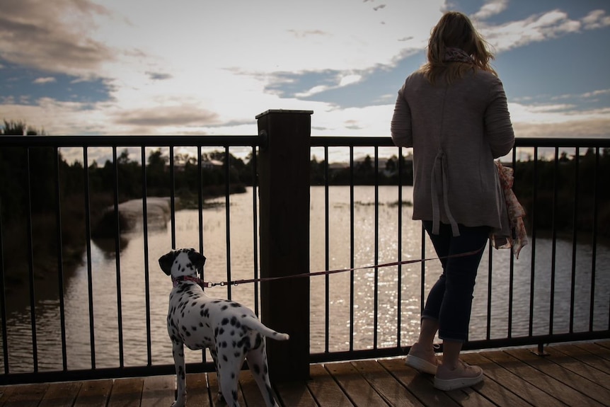 A blonde woman looks out over a river, with her dalmation on a lead beside her.