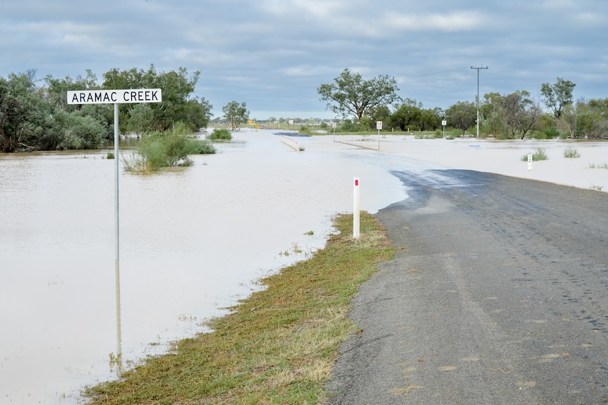 A white sign says Aramac Creek, as floodwaters cover a bridge in the background.