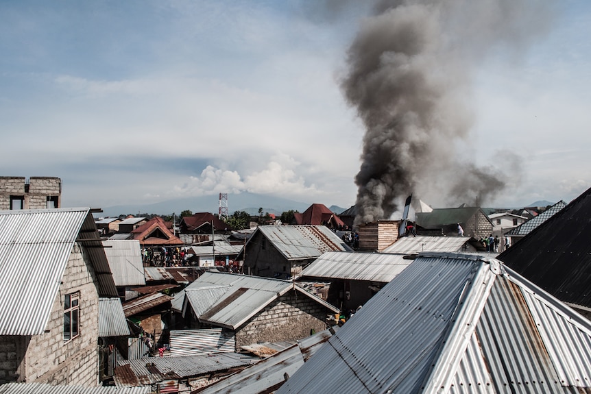 A panoramic image of corrugated iron houses shows smoke billowing on the horizon.
