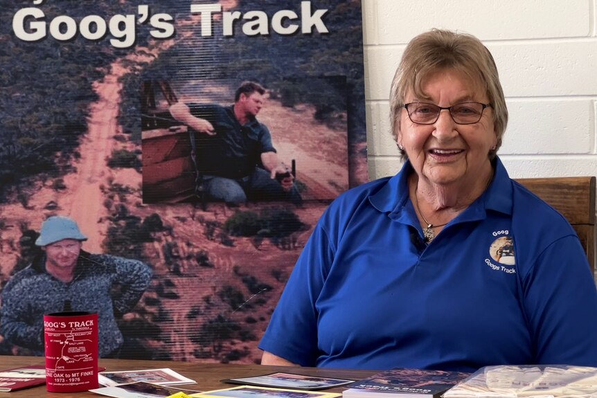 Elderly woman in blue shirt sitting at table with Googs Track poster in background