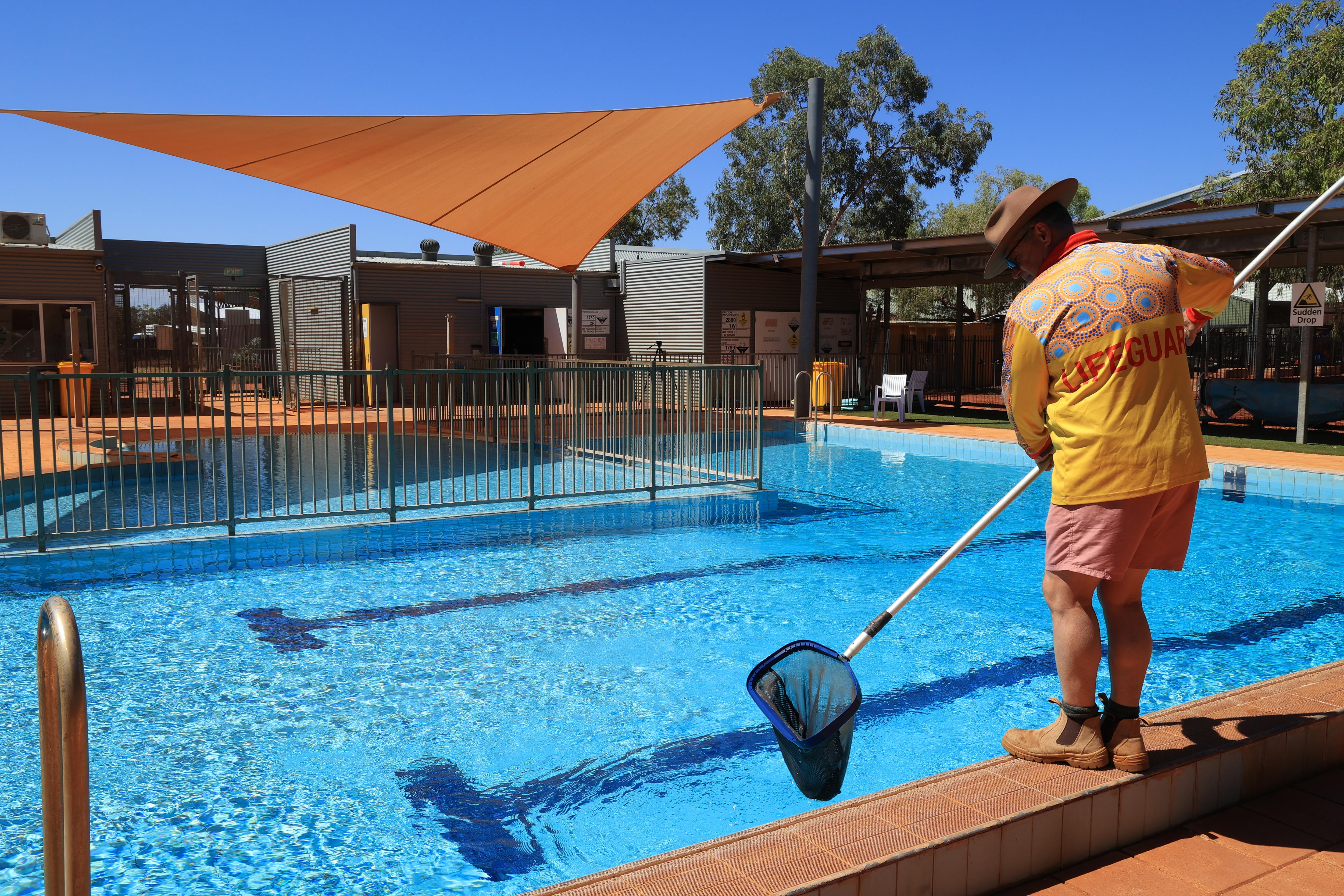 A lifeguard stands on the edge of a blue community pool, holding a net used to scoop leaves from the water.