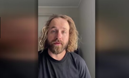 A man with shoulder-length blonde hair looking into the camera as he speaks