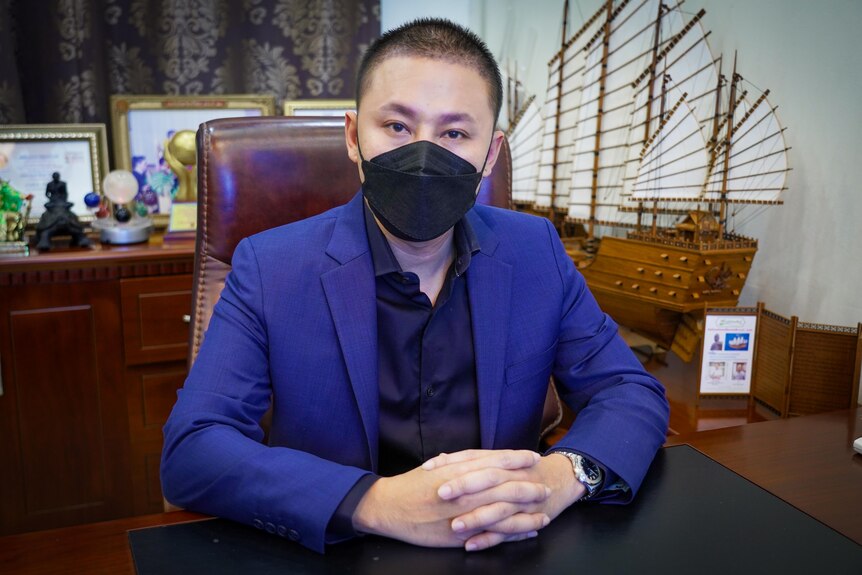 Un Thaïlandais vêtu d'un costume bleu et d'un masque noir est assis à un bureau, les mains jointes